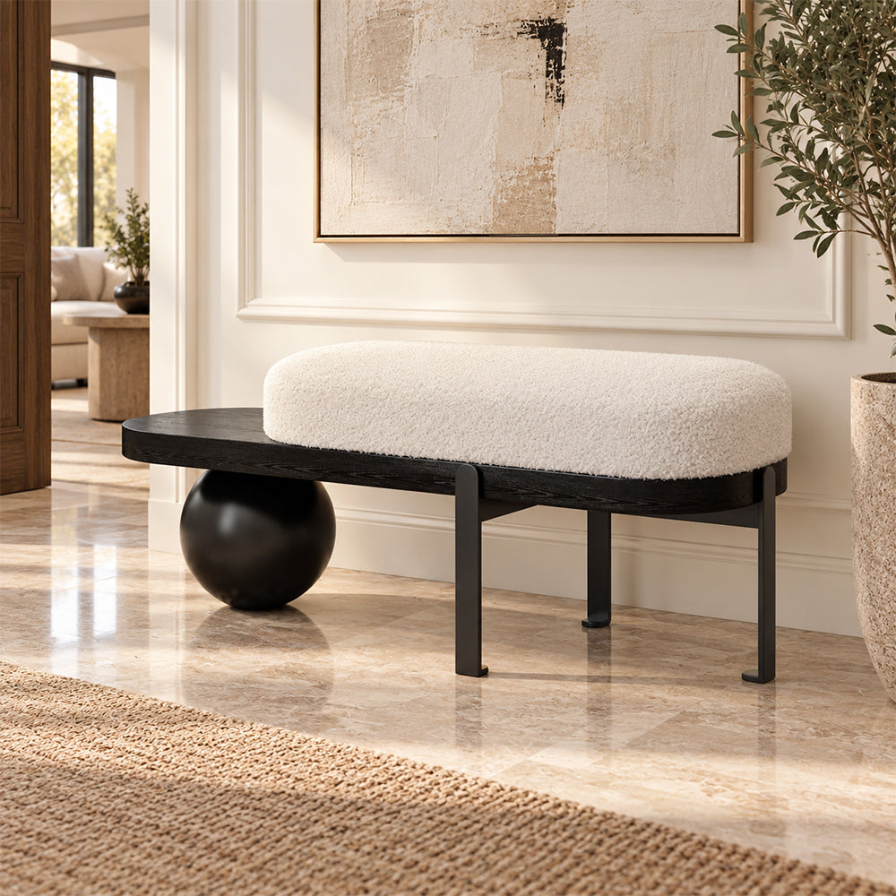 Cream upholstered entryway bench with a black frame, exposed side shelf and black spherical support.