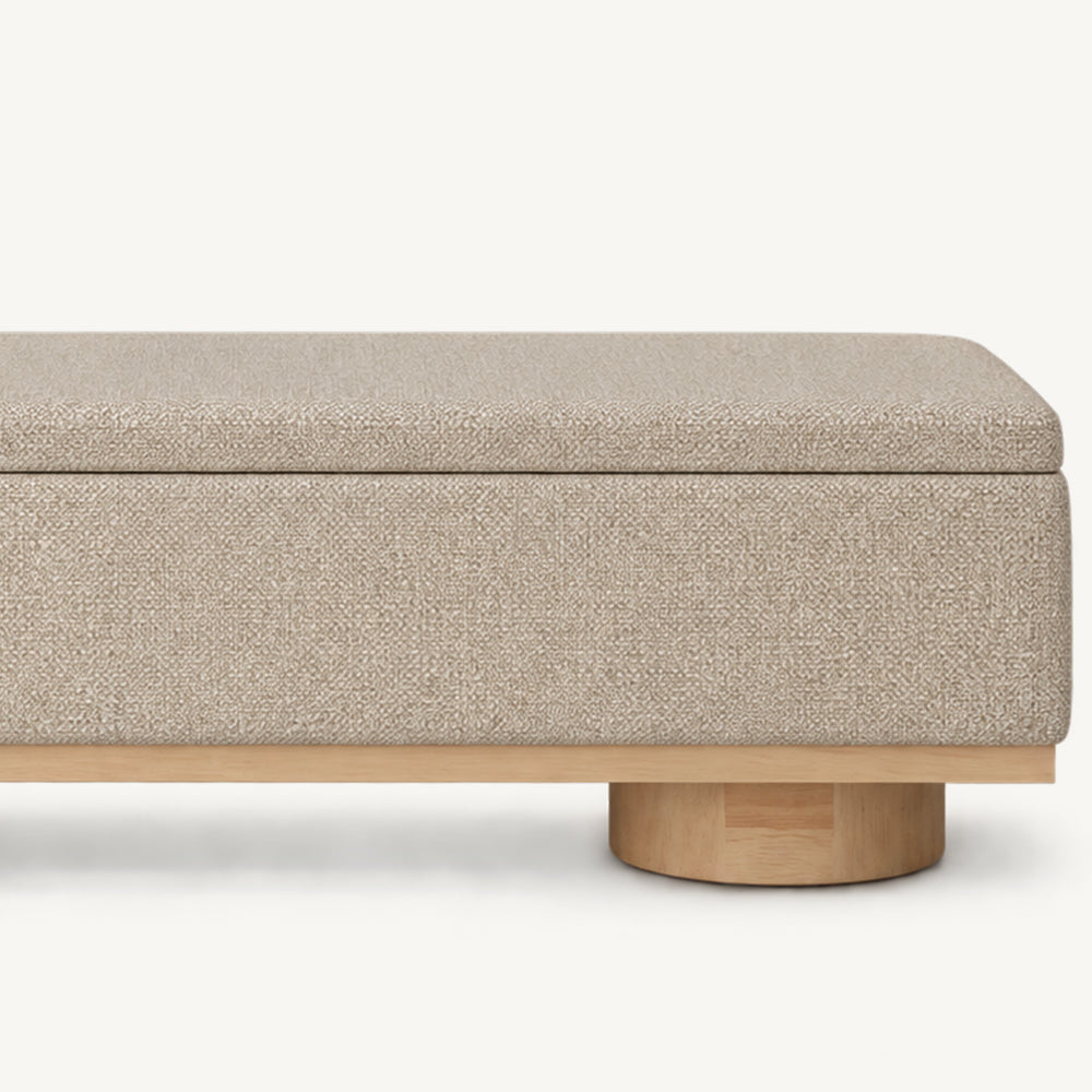 Front detail of the beige bench's lid seam, textured upholstery and wood-tone base trim.