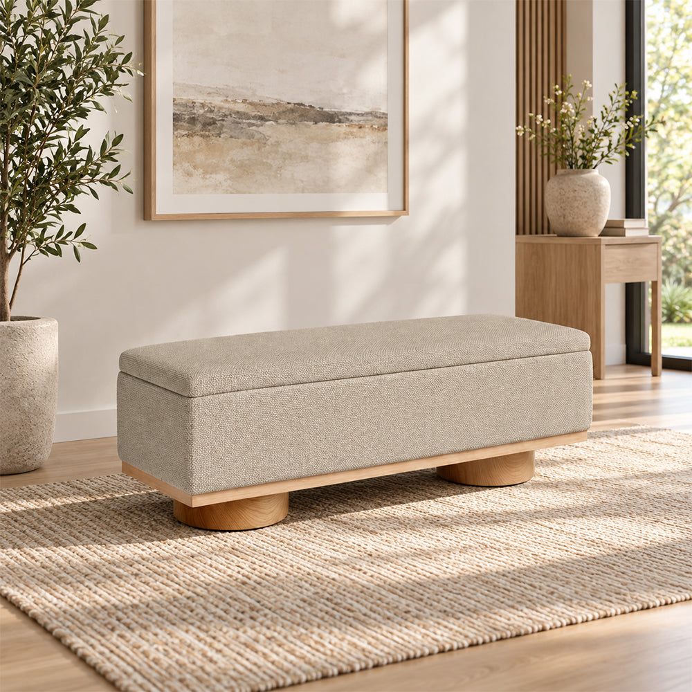 Angled room view of the beige upholstered bench showing its padded lid and round wooden-tone supports.