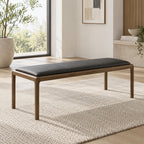 Black padded bench with a slim brown frame and four slender legs beside a large window.
