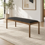 Black upholstered bench with a thin wood-tone frame and slender legs in a sunlit room.