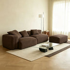 Angled view of the brown modular sofa with rounded arms and a matching ottoman beside a window.