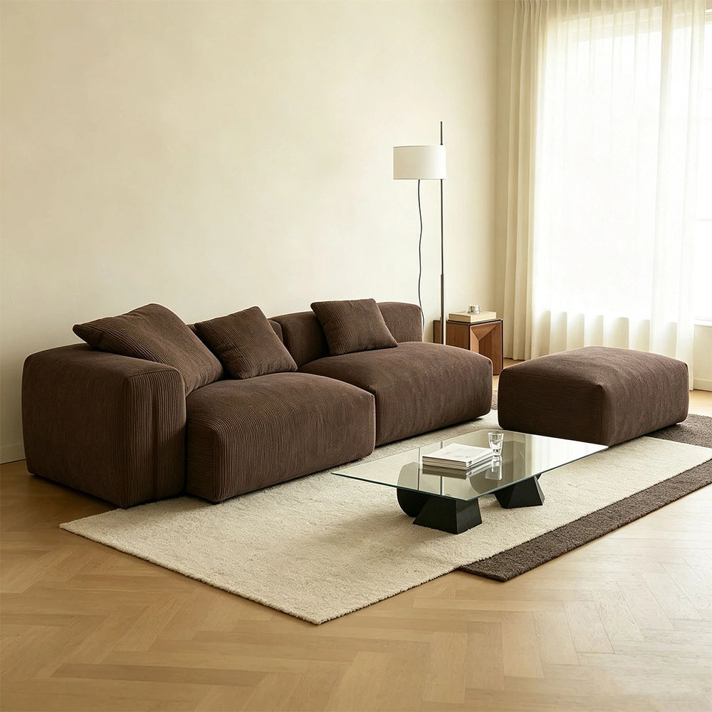 Angled view of the brown modular sofa with rounded arms and a matching ottoman beside a window.