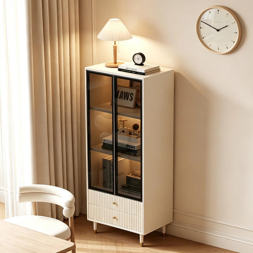 Angled room view of the cream glass display cabinet with fluted drawers and gold-tone feet.