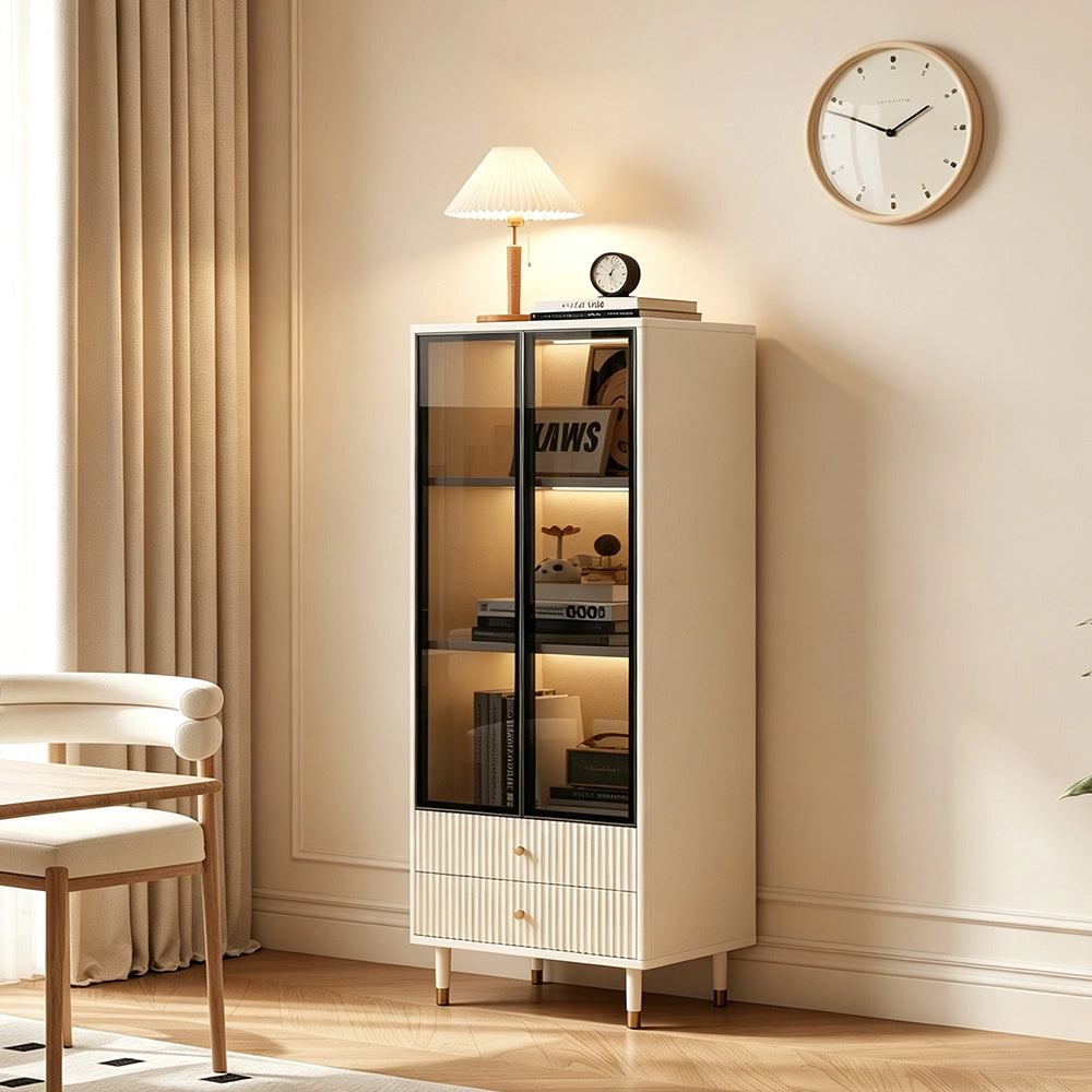 Cream glass display cabinet standing beside a chair, with illuminated shelves holding books and ornaments.