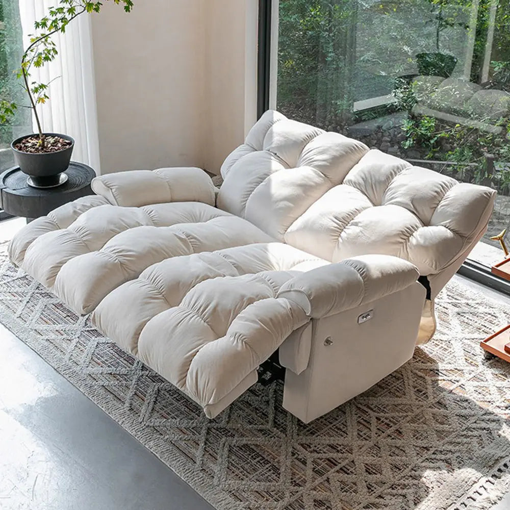 Cream loveseat with both backrests lowered and footrests extended beside a large window.