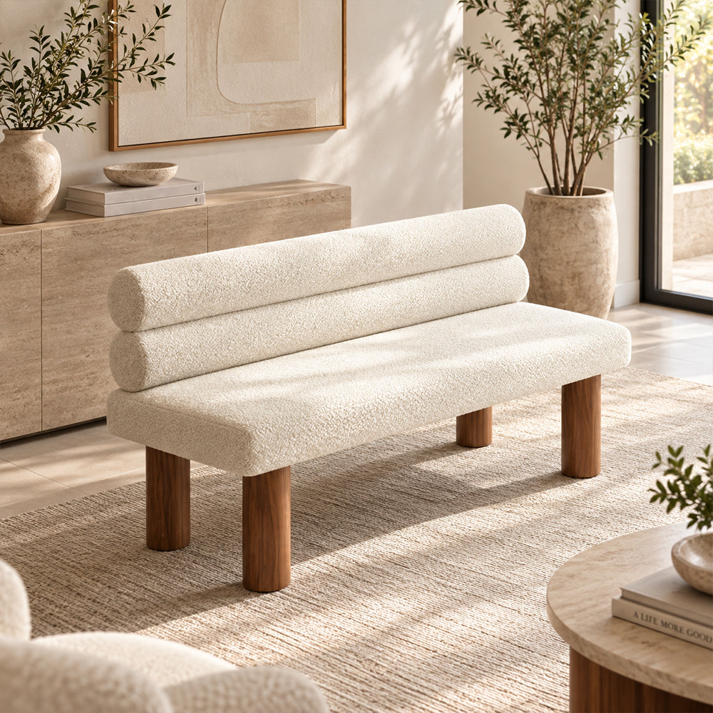 Cream textured bench with two cylindrical back cushions and brown round legs in a bright living room.