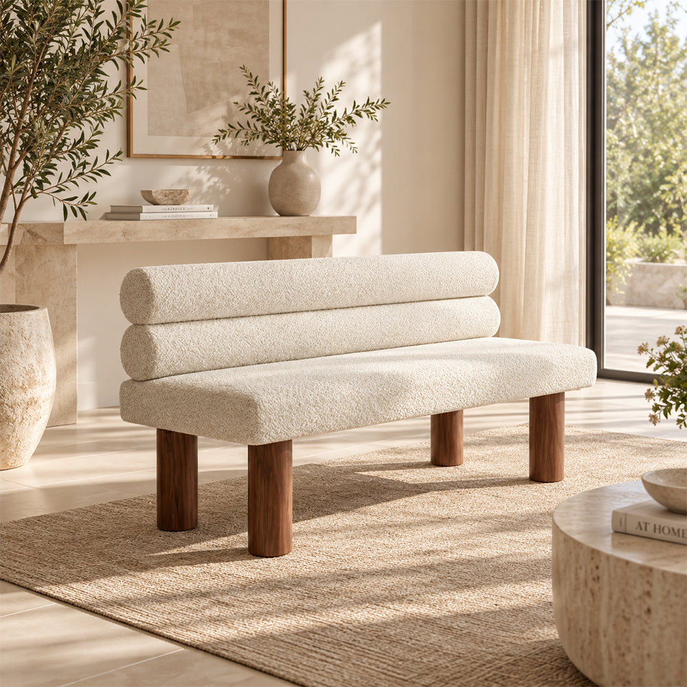 Angled view of the cream double-roll-back bench beside a window and low tables.