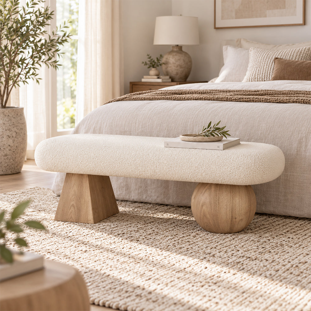 Cream upholstered bench with light wood-tone geometric supports at the foot of a bed.