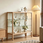 Light wood-tone display cabinet with clear glass doors and shelves holding white ornaments and flowers.