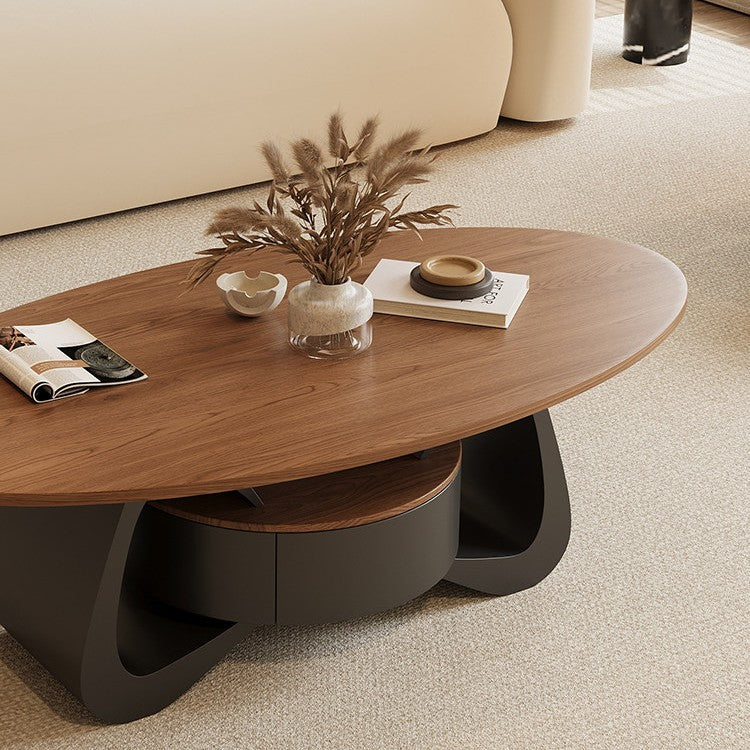 Oval wood-tone coffee table with a dark curved storage base, styled with a vase and books beside a cream sofa.
