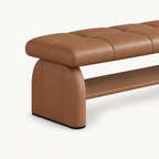 Angled detail of the brown bench's rounded upholstered side support, stitched cushion and lower shelf.