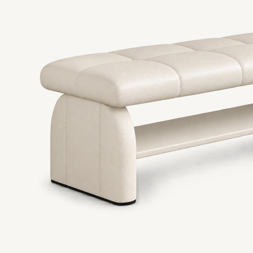 Angled detail of the cream bench's rounded upholstered side support, stitched cushion and lower shelf.