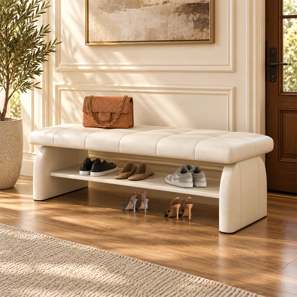 cream padded shoe bench with a stitched seat, broad upholstered supports and a lower shelf holding shoes.