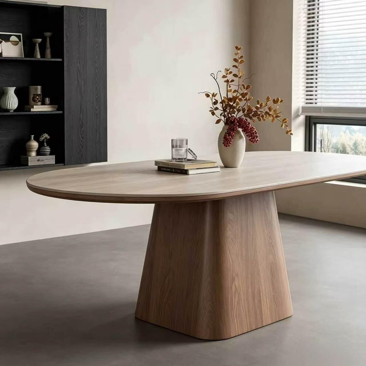 Pale oval dining table with a brown tapered central pedestal, styled with a vase and glass.