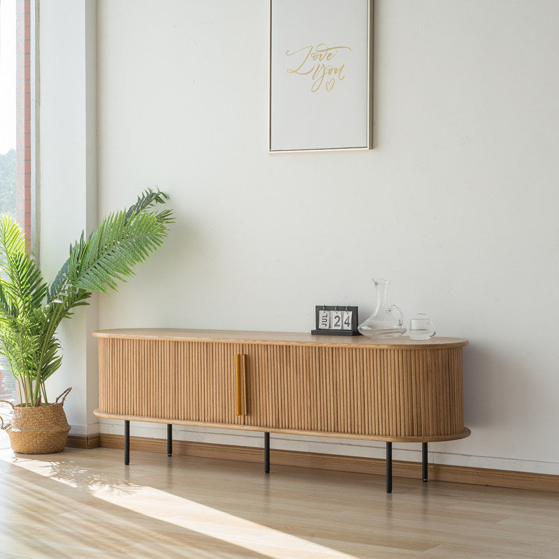 Angled view of the rounded slatted-door TV stand on thin black legs beside a plant.