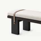 Detail of the cream upholstered bench's left end, brown strap and broad black legs.