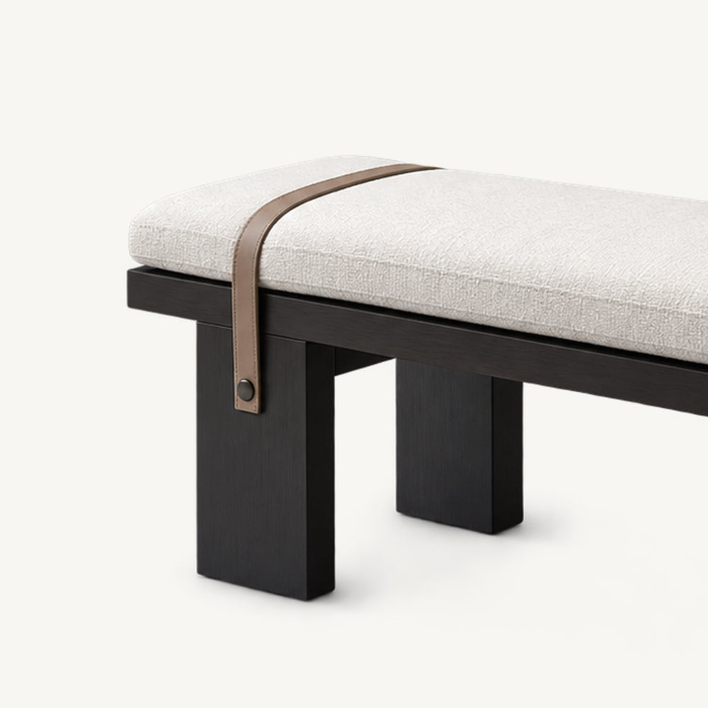 Detail of the cream upholstered bench's left end, brown strap and broad black legs.