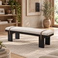 Cream upholstered bench with a black block-leg frame and two brown straps over the cushion.