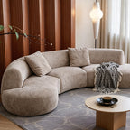 Taupe curved modular sofa beside small round tables, showing its rounded arm and textured upholstery.